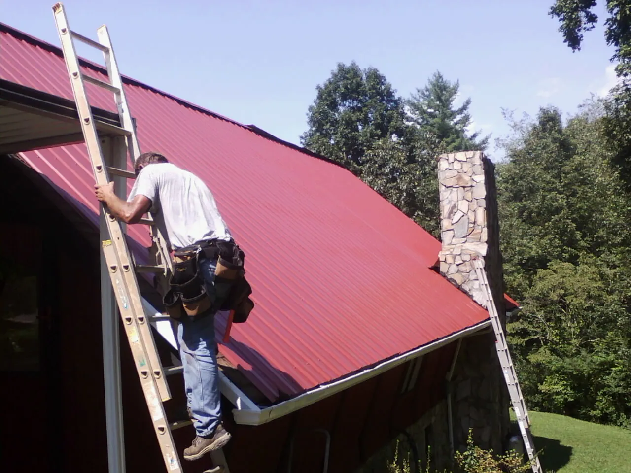 Roofer on a ladder beside a red metal roof and stone chimney — Soffit & Fascia Repair by Roofing Brothers in Laporte